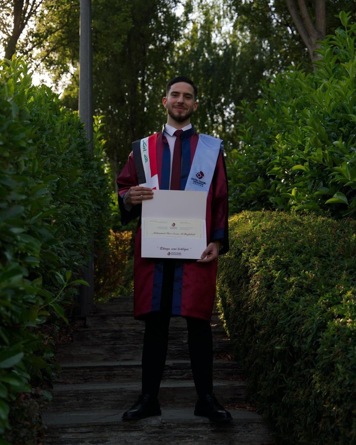 Mohammed at his graduation in a burgundy gown with an Iraqi flag stole, holding his diploma
